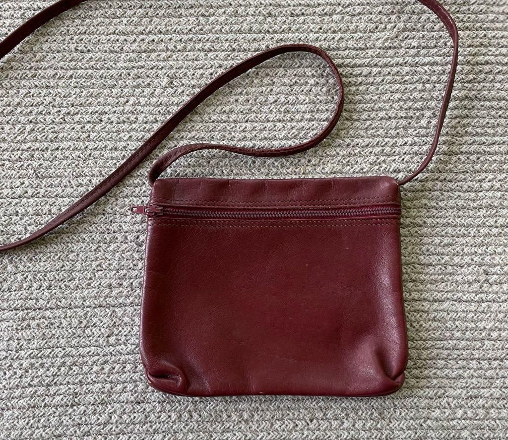Compact oxblood leather bag with a long strap against a neutral woven background.