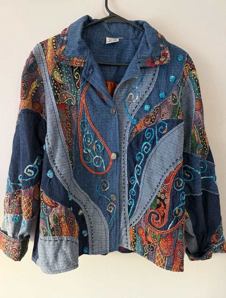 Dark denim jacket with colorful botanical embroidery and patchwork panels, hanging against a light wall.