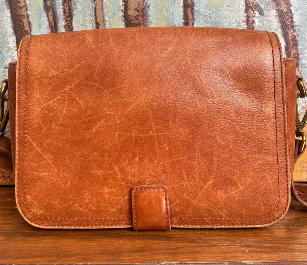 Close-up of a cognac-brown leather flap shoulder bag with natural creases and understated brass hardware.