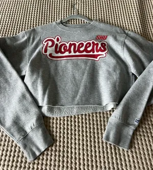 Show your school spirit with this Pioneers gray cropped sweatshirt. Soft gray fabric with bold red Pioneers lettering. Comfortable crewneck and relaxed fit for casual wear. Easy to pair with jeans or leggings for a laid-back look. thumbnail 1