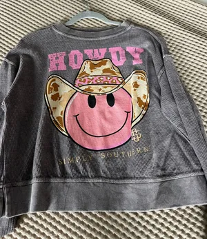 Gray crewneck pullover featuring a large pink smiley wearing a cow-print cowboy hat. "HOWDY" text in pink above the graphic. Casual, playful design from Simply Southern. thumbnail 1