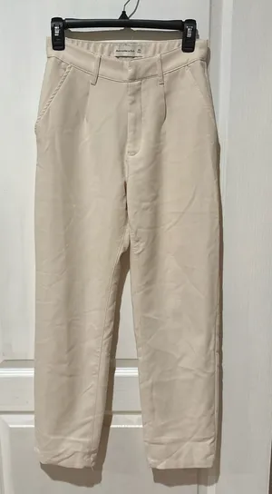 Abercrombie & Fitch high-waisted trousers. The design features a classic straight-leg silhouette with subtle front pleating, diagonal side pockets, and a button-and-zip fly closure in a cream or "oyster" shade. thumbnail 1