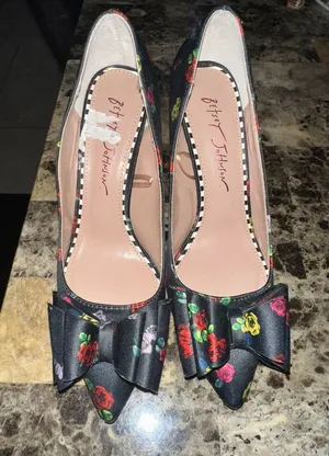 These are Betsey Johnson 'Bret' pumps featuring a black satin upper with a colorful floral print and a prominent bow detail on the pointed toe. thumbnail 1