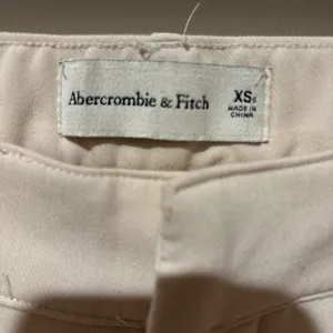 Abercrombie & Fitch high-waisted trousers. The design features a classic straight-leg silhouette with subtle front pleating, diagonal side pockets, and a button-and-zip fly closure in a cream or "oyster" shade. thumbnail 3