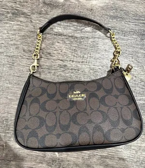 Elegant Coach shoulder bag in the classic brown and black Signature “C” canvas. Features black trim, gold-tone hardware, a zip-top closure, and a stylish black shoulder strap with decorative gold chain details. Compact yet roomy—perfect for everyday essentials or a chic night out. thumbnail 1